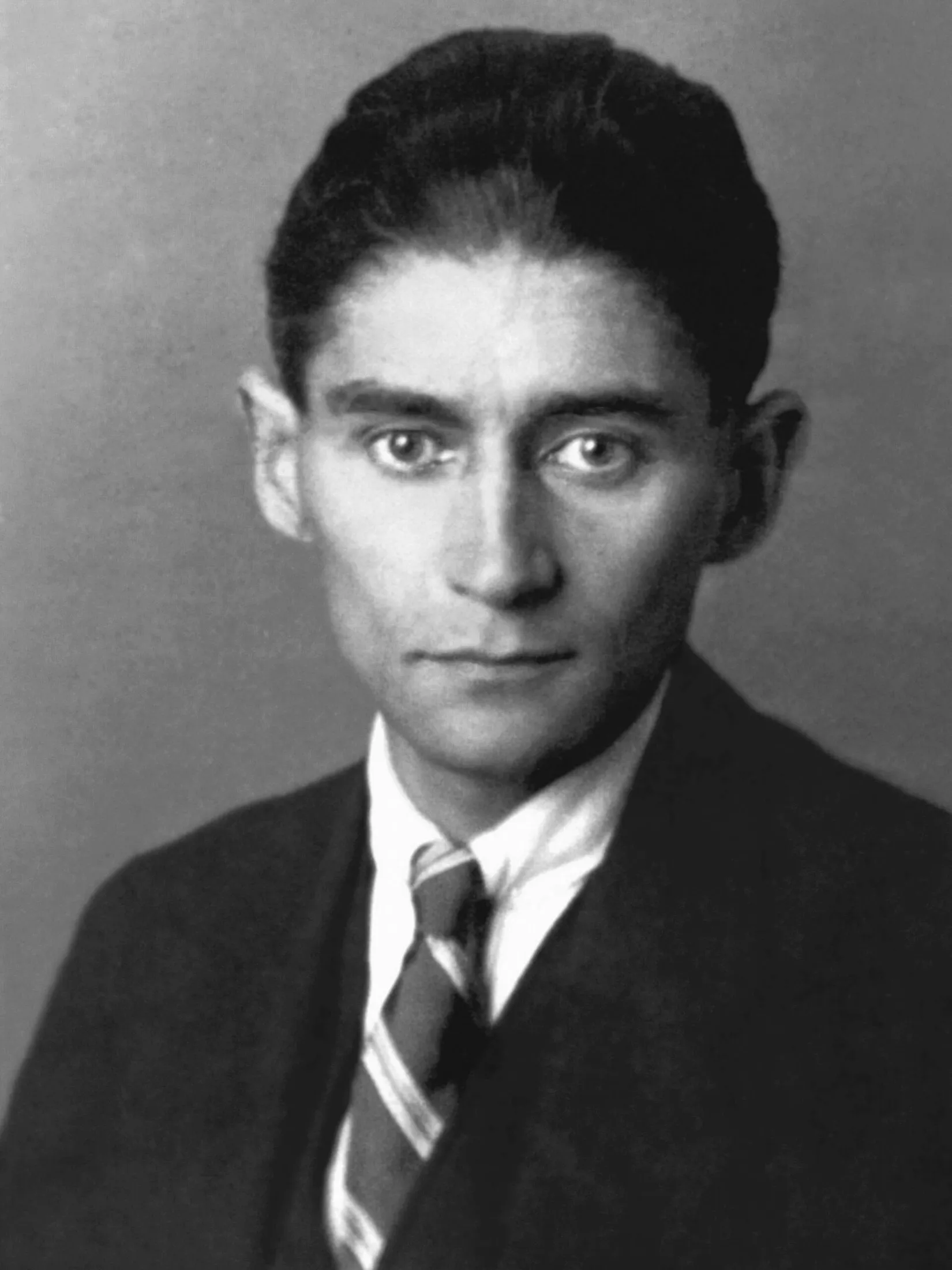 A studio portrait of a thin man in a dark suit and tie with short dark hair and large ears, looking straight at the camera.