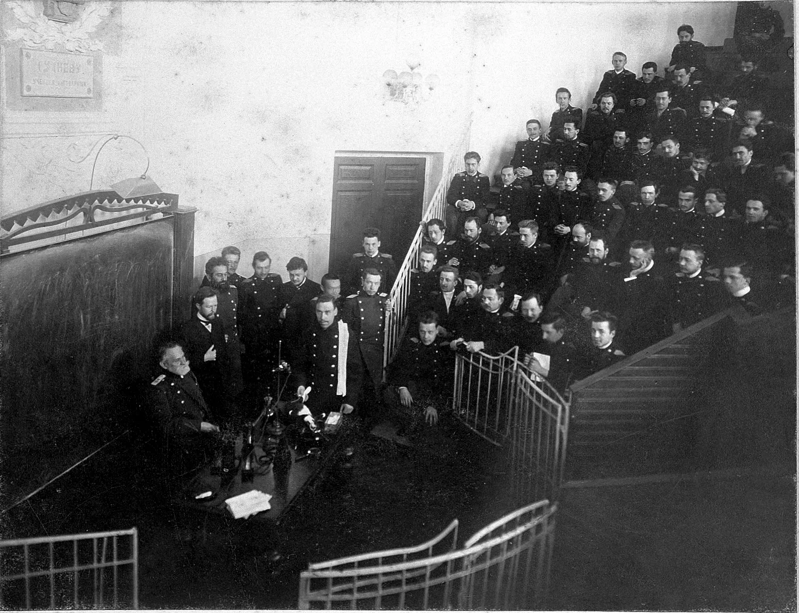 A lecture theatre full of men in uniform and white coats, watching an elderly bearded man at a table with a dog on it.