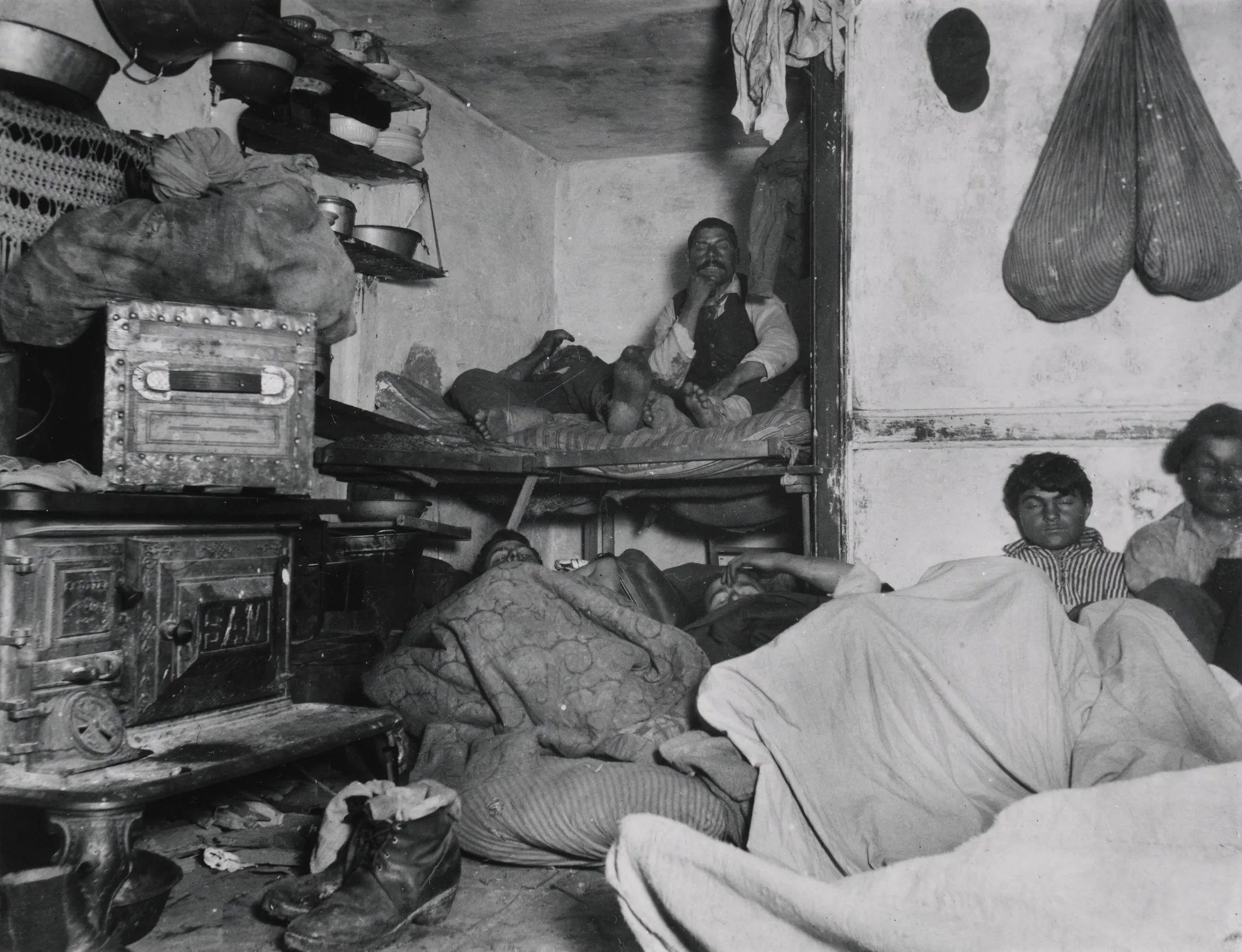 A cramped tenement room at night, seven men lying and sitting on bunks, boxes and the floor, with clothes and pots hung on the walls.