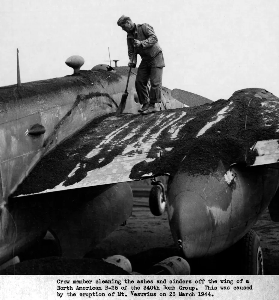 A man in uniform stands on the wing of a twin-engined bomber, sweeping off a thick layer of grey ash.